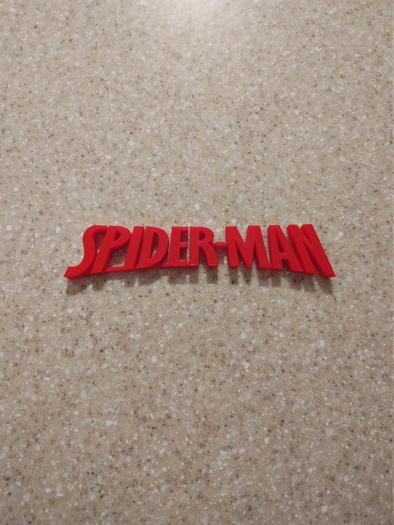 May include: Red 3D plastic letters spelling out "SPIDER-MAN" on a beige speckled surface.