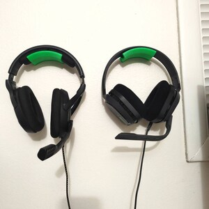 May include: Two black gaming headsets with green accents are hanging on a white wall. The headsets have a microphone attached to each one.