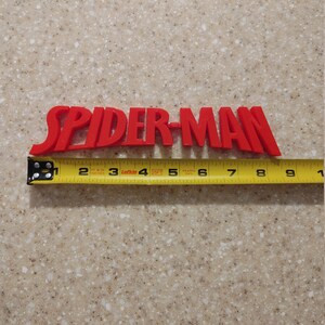 May include: Red plastic "SPIDER-MAN" lettering with a yellow measuring tape underneath. The tape measures 23 cm long.