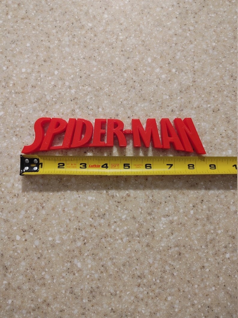 May include: Red plastic "SPIDER-MAN" lettering with a yellow measuring tape underneath. The tape measures 23 cm long.