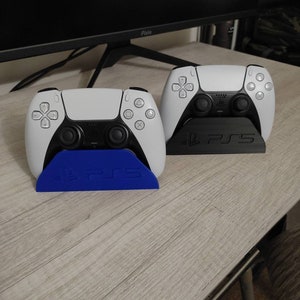 Op de afbeelding: Twee witte PlayStation 5-controllers worden getoond op hun standaards. De standaards zijn zwart met het PlayStation-logo en de woorden "PS5" in wit. De standaard aan de linkerkant is blauw.