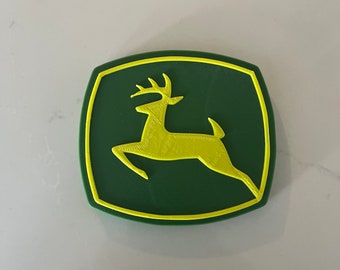 Puede incluir: Un logotipo impreso en 3D de un ciervo saltando, en verde y amarillo. El ciervo es amarillo y el fondo es verde.