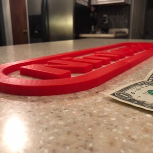 Pode incluir: Uma placa vermelha impressa em 3D que diz "WINNING" em letras maiúsculas. A placa tem formato oval. Uma nota de um dólar está ao lado da placa em uma bancada salpicada.