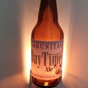 May include: A brown glass beer bottle night light with a white label that reads "Lagunitas DayTime Ale Fractional IPA" and features a cartoon dog. The bottle is plugged into a white electrical outlet.