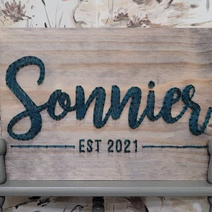 Può includere: Un cartello di legno con la parola "Sonnies" scritta in filo blu turchese. Il cartello include anche il testo "EST 2021" sotto il nome.