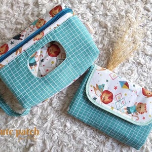 Pode incluir: Uma bolsa de tecido com padrão de grade azul-petróleo, zíper e abertura retangular, acompanhada por uma carteira dobrável combinando. Ambos os itens apresentam uma estampa branca com leões de desenho animado, pipas e amarelinhas.