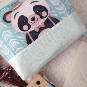 Pode incluir: Uma bolsa azul clara com zíper com uma ilustração de um urso panda usando um chapéu e uma gravata borboleta. O panda está em um padrão de chevron azul claro. A parte inferior da bolsa tem um design tecido verde claro.
