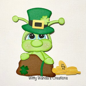 May include: A green leprechaun-like creature wearing a green hat with a gold buckle and shamrock. It holds a brown bag with a shamrock and gold coins. The text "Witty Wanda's Creations" is at the bottom.