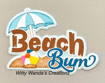 Puede incluir: Una decoración de papel troquelado en capas con el texto Beach Bum en marrón y azul, una sombrilla de playa a rayas azul claro y blanco, y una pelota de playa colorida sobre una base de arena. El texto Witty Wanda's Creations está abajo.