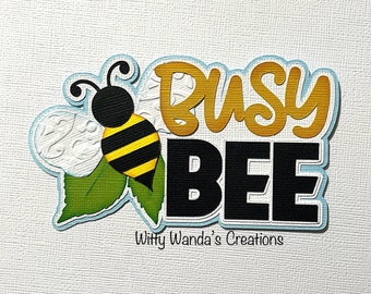 Puede incluir: Una abeja negra y amarilla con una hoja blanca y verde detrás. El texto "Busy Bee" está en letras doradas y negras con un contorno azul.