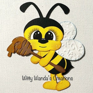 May include: A die-cut paper craft of a cartoon bee with yellow and black stripes, large eyes with long lashes, and white textured wings. The bee holds a brown wooden honey dipper with glossy amber honey. Text reads Witty Wanda's Creations.