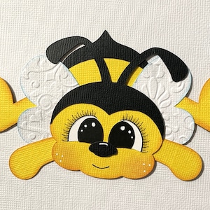 May include: A yellow and black paper cutout of a bee with large eyes and a smiling mouth. The bee has white wings with a decorative pattern.