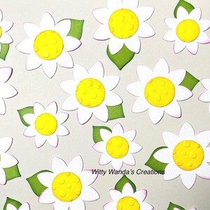 May include: White daisy cutouts with yellow centers and green leaves are scattered across a light background. The daisy petals have a pink outline. The text "Witty Wanda's Creations" is visible in the lower right corner. These are likely craft supplies.