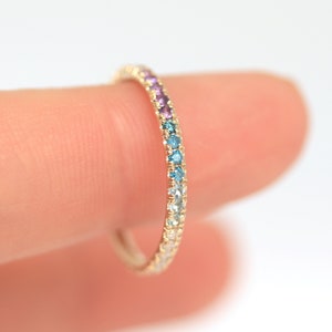 May include: A gold band ring with a rainbow of small gemstones set in the band. The gemstones are purple, blue, and clear.