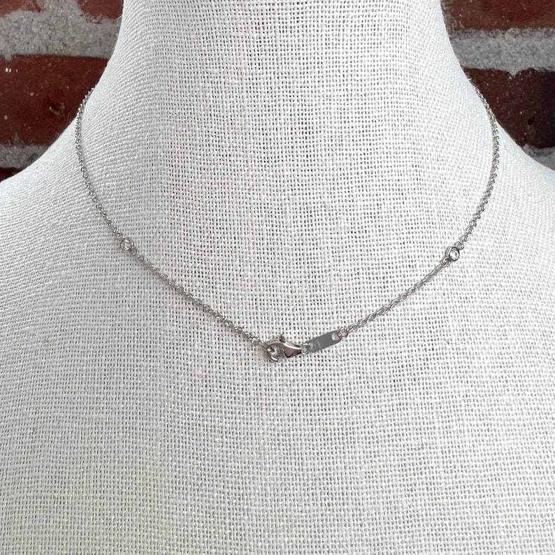 May include: A delicate silver necklace with small, round embellishments. The necklace is displayed on a white mannequin, showcasing its fine chain and clasp. The necklace is a simple, elegant piece of jewelry.
