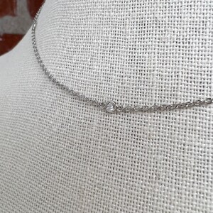 May include: A delicate silver necklace with a small, round, clear gemstone pendant. The chain is thin and has a subtle, textured design. The necklace is displayed on a neutral-colored fabric background.