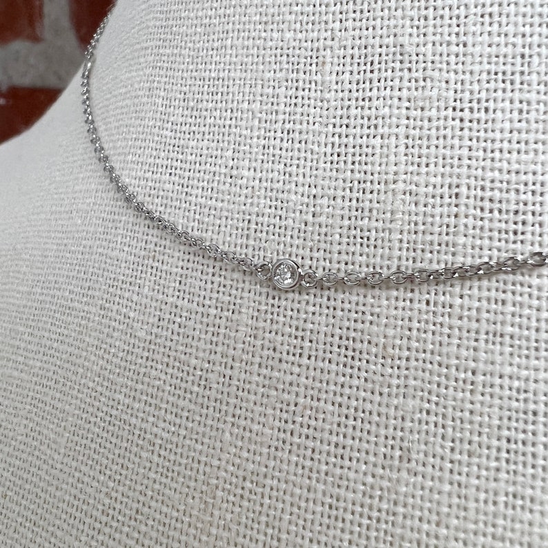 May include: A delicate silver necklace with a small, round, clear gemstone pendant. The chain is thin and has a subtle, textured design. The necklace is displayed on a neutral-colored fabric background.