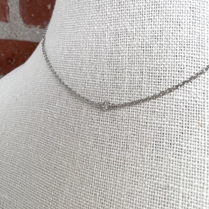 May include: A delicate silver necklace with a small, round, clear gemstone pendant. The necklace is displayed on a white fabric background, showcasing its simple elegance. The chain is thin and the pendant is centered.