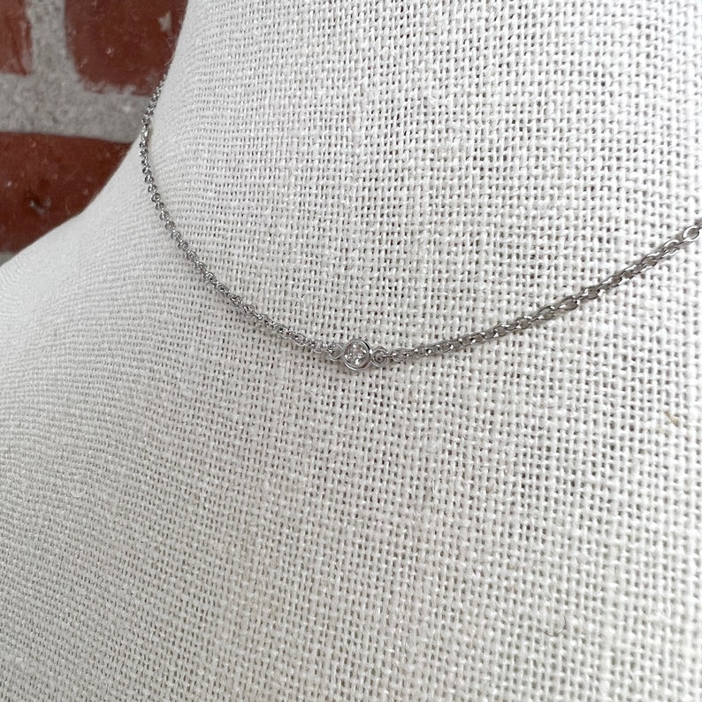 May include: A delicate silver necklace with a small, round, clear gemstone pendant. The necklace is displayed on a white fabric background, showcasing its simple elegance. The chain is thin and the pendant is centered.