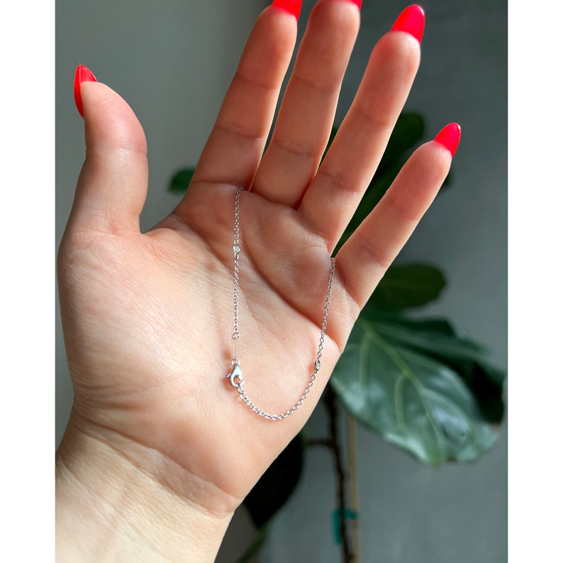 May include: A delicate silver necklace with a small chain and a lobster clasp. The necklace features several small, round embellishments. The jewelry is displayed against a neutral background.