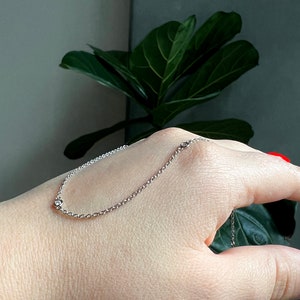 May include: A delicate silver chain bracelet with three small, clear gemstones. The bracelet is displayed on a hand, with a green plant in the background. The bracelet is a simple, elegant piece of jewelry.