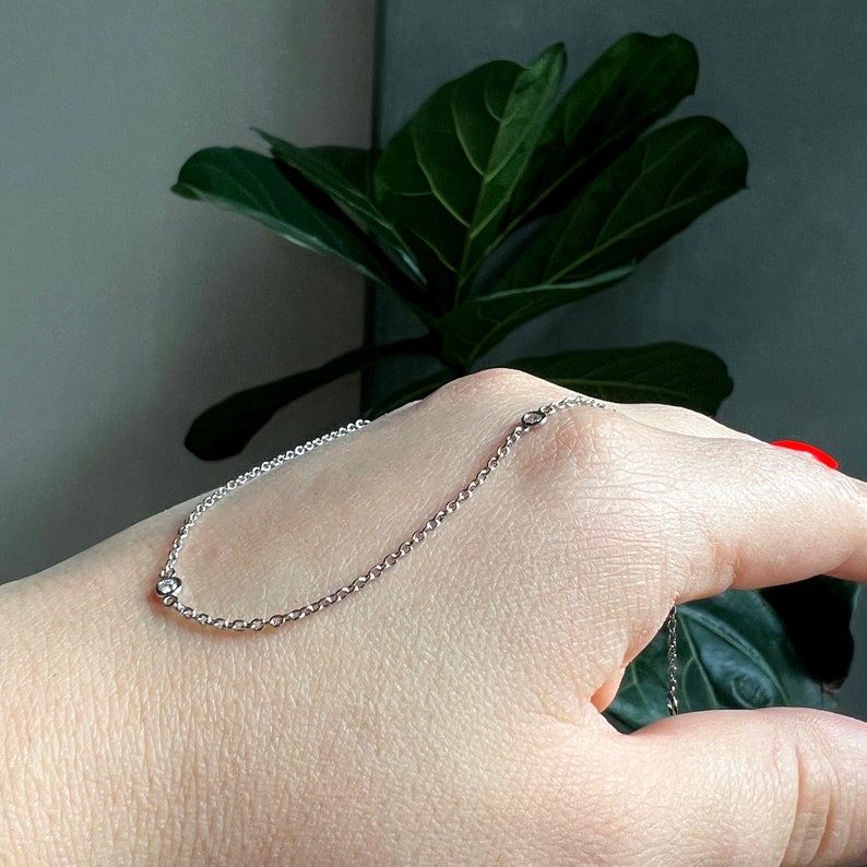 May include: A delicate silver chain bracelet with three small, clear gemstones. The bracelet is displayed on a hand, with a green plant in the background. The bracelet is a simple, elegant piece of jewelry.
