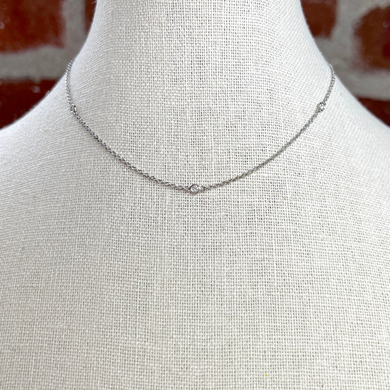May include: A delicate silver necklace with three small, round, clear gemstones evenly spaced along the chain. The necklace is displayed on a white mannequin, with a brick wall in the background.