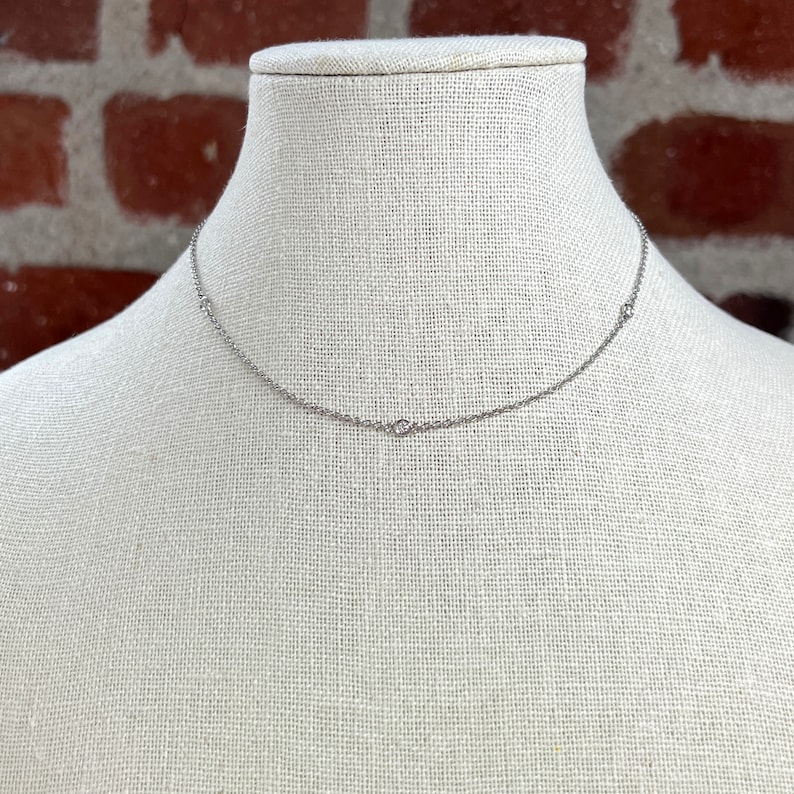 May include: A delicate silver necklace with three small, round embellishments. The necklace is displayed on a mannequin with a neutral-colored fabric background, set against a brick wall.