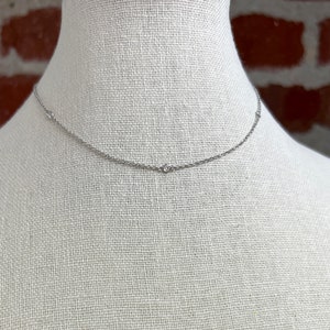 May include: A delicate silver necklace with small, sparkling gemstones evenly spaced along the chain. The necklace is displayed on a white mannequin, with a brick wall in the background. This minimalist necklace is a classic jewelry piece.