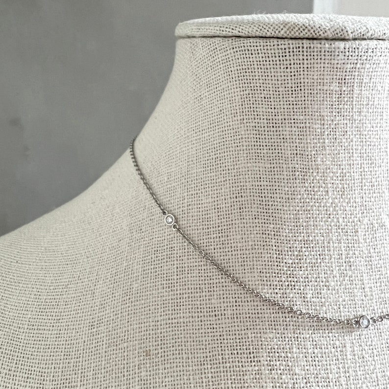 May include: A delicate silver necklace with three small, round, clear gemstones evenly spaced along the chain. The necklace is displayed on a neutral-colored mannequin, showcasing its simple elegance and design.