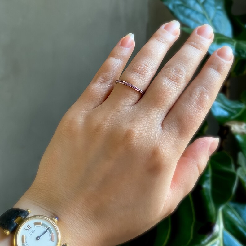 May include: A delicate gold band ring with a row of small, deep red gemstones. The ring is worn on the ring finger of a hand.