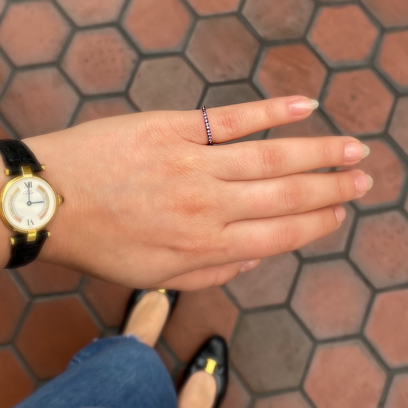 May include: A delicate gold band ring with a row of small pink gemstones. The ring is worn on the index finger of a hand.