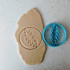 May include: A blue plastic cookie cutter in the shape of a lightning bolt, used to cut out a cookie in the shape of the Grateful Dead logo. The cookie cutter is next to a piece of cookie dough with the logo cut out.
