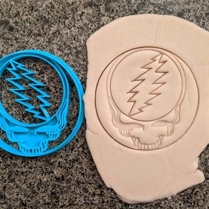 May include: A blue plastic cookie cutter in the shape of the Grateful Dead's Steal Your Face logo. The cutter is next to a piece of dough with the logo imprinted in it.