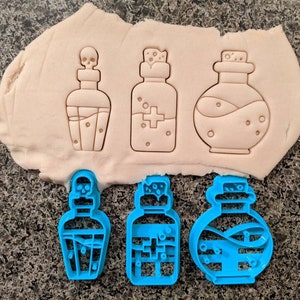 May include: Three potion bottle cookie cutters in blue, next to three potion bottle shapes pressed into dough. The potion bottle shapes include a skull, a cross, and a wavy design. The dough is light brown, and the background is a speckled gray.