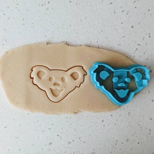 May include: A blue plastic cookie cutter in the shape of a bear's head with a smiling face. The cutter is on top of a piece of cookie dough.
