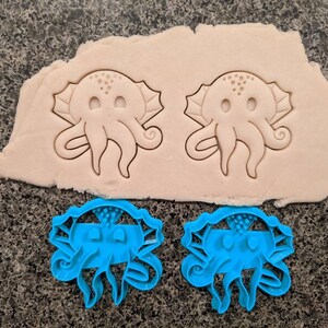 Puede incluir: Dos cortadores de galletas de plástico azul con forma de pulpos de dibujos animados con ojos grandes y una sola tentacula rizada. Los cortadores están sobre una superficie marrón claro. Dos cortadores de galletas blancos con forma del mismo pulpo de dibujos animados están sobre una superficie marrón claro.