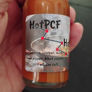 May include: A clear glass bottle filled with orange-red hot sauce. The label features the text HotPCF and a graphic of a wok with steam. The ingredients listed are Tabasco, vinegar, miso, red pepper, black pepper, chili oil, and sea salt.