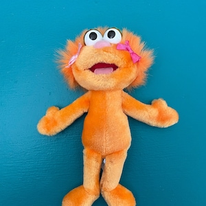 Puede incluir: Juguete de peluche naranja con una gran sonrisa y ojos grandes. El juguete tiene lazos rosas en la cabeza y está acostado sobre una superficie azul.