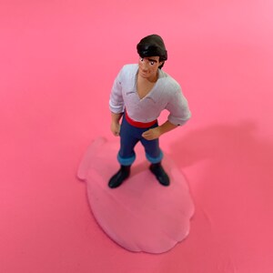Puede incluir: Una figura de plástico de un príncipe de Disney, el príncipe Eric de La Sirenita, que lleva una camisa blanca, pantalones azules y botas negras. Está de pie sobre una base rosa.