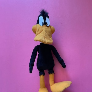 Puede incluir: Un juguete de peluche negro y amarillo de Daffy Duck de la caricatura de Looney Tunes. El juguete está de pie sobre un fondo rosa.