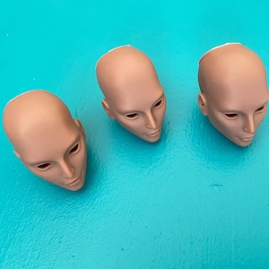 Può includere: Tre teste di bambola non dipinte con tratti del viso dettagliati, inclusi occhi chiusi e un'espressione neutra. Le teste sono di colore marrone chiaro e sono disposte su una superficie turchese brillante.