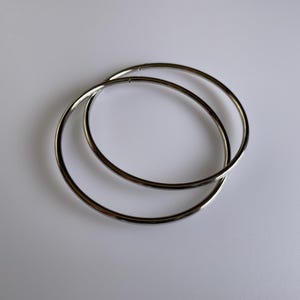 May include: Two silver hoop earrings, one slightly larger than the other, are lying on a white surface.