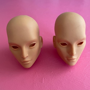 Può includere: Due teste di bambola non dipinte, con orbite oculari aperte e lineamenti delicati. Le teste sono posizionate su uno sfondo rosa acceso, evidenziando le loro superfici lisce e vuote. Le teste di bambola sono di colore beige chiaro.