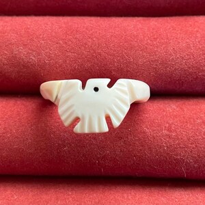 May include: A ring featuring a carved off-white bird motif with a small black dot representing an eye. The bird design is centered on the band, with wings extending outward and a tail section pointing downward, creating a singular focal point.