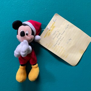 Puede incluir: Un Mickey Mouse de peluche rojo y blanco con un gorro de Papá Noel. El juguete de peluche es un clip y tiene una etiqueta que dice "PROTOTY SAMPLE SOFT GOODS", "Monica F", "54269", "Kimas Mickey & Minnie Clip On", "2001 Amas" y "Cu 1/100".