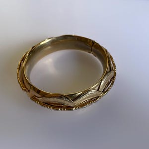 May include: A gold bangle bracelet with a decorative design of overlapping leaves. The bracelet is made of two different shades of gold.