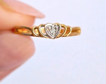 May include: A gold ring with a heart-shaped design. The ring features a central heart-shaped setting with a cluster of small, clear stones. The band is a simple, polished gold. The ring is held against a plain white background.
