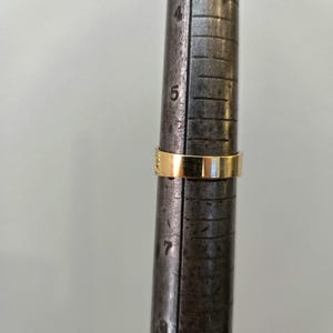 May include: A gold band ring is positioned on a metal ring mandrel. The ring rests between the size 5 and 6 markings on the tool, which features engraved numbers and vertical lines to indicate ring size measurements in inches.
