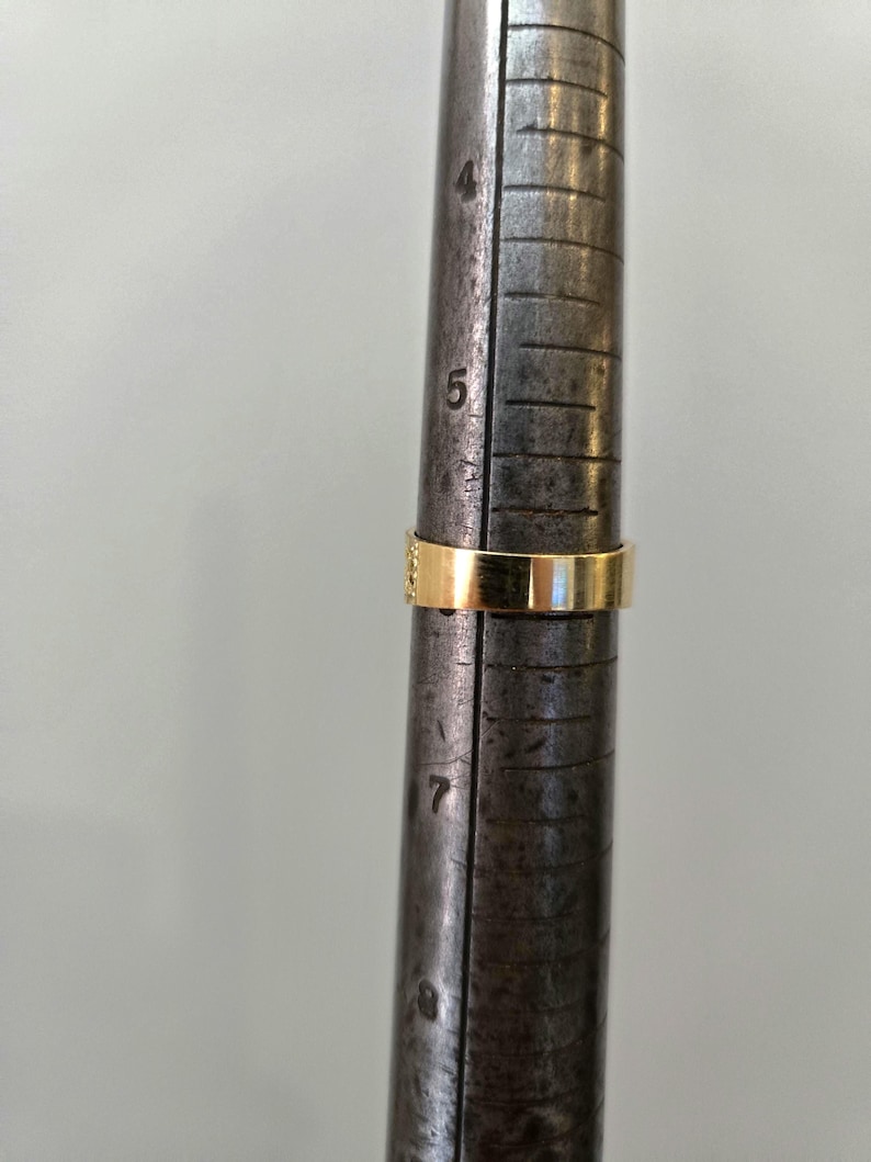 May include: A gold band ring is positioned on a metal ring mandrel. The ring rests between the size 5 and 6 markings on the tool, which features engraved numbers and vertical lines to indicate ring size measurements in inches.